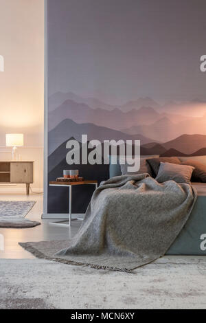 Grey blanket and pillows on bed against mountain wallpaper in cozy bedroom interior with light Stock Photo