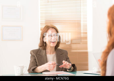 Smiling professional advisor in white glasses sitting in her office behind a desk and talking to a woman