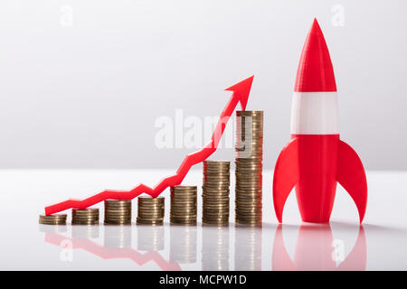 Close-up Of Red Rocket Besides Stacked Coins And Arrow Showing Upward Direction Stock Photo