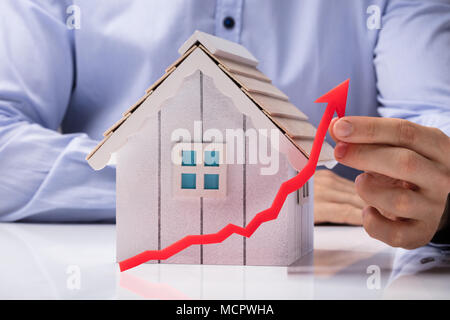 Close-up Of A Person's Hand Holding Arrow In Front Of House Model On White Desk Stock Photo