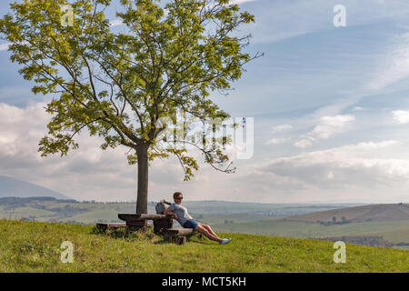 Caucasian middle aged woman hiker have a rest on wooden bench. Summer hills landscape close to Liptovsky Trnovec village in Slovakia. Stock Photo