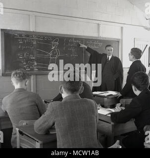 1950s, historical picture of an adult male teacher in a shirt and tie and wearing a traditional gown using a large blackboard to teach or educate a small group of school pupils sitting at desks, England, UK. Stock Photo