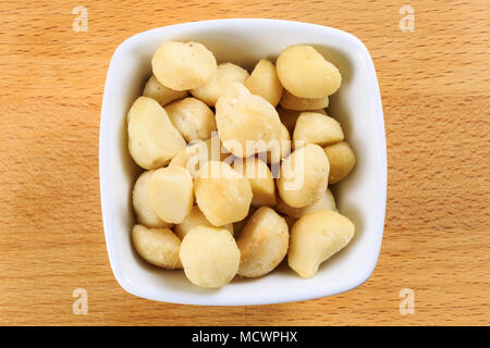 Overhead shot of raw macadamia nuts in a white bowl on a wooden board background. Macadamia nuts are the seeds of the macadamia tree, Macadamia tetrap Stock Photo