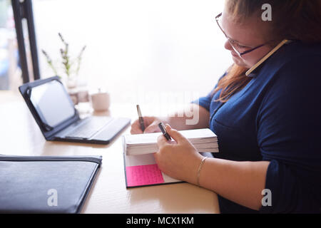 Moderately overweight caucasian woman seated at her desk busy multitasking while she writes in her notebook and talks on her phone, that is pinched between her head and shoulders. Stock Photo