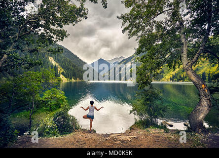 Young woman is practicing yoga tree pose at mountain lake with overcast sky background Stock Photo