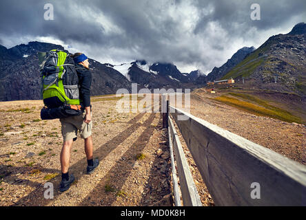 Hiker in with big green backpack walking to the snowy mountains at overcast dark sky background in Shymbulak Ski Resort in Kazakhstan Stock Photo