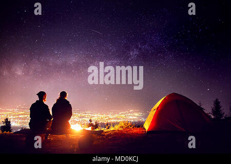 Happy couple in silhouette sitting near campfire and orange tent. Night sky with Milky Way stars and city lights at background. Stock Photo