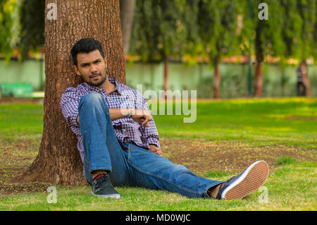 Young stylish happy man sitting on the grass leaning against tree Stock Photo