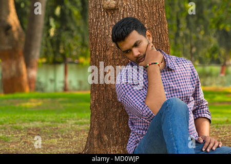 Young guy sitting and thinking leaning against tree Stock Photo