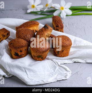 small round baked muffins with dry fruits and raisins on a white textile napkin, gray background, behind a bouquet of daffodils Stock Photo