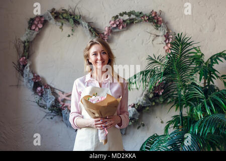 Image of smiling florist with bouquet in hands against background of scenery from flowers in form of heart on wall Stock Photo