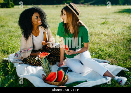 Two pretty charming multi-race girlfriends with natural make-up and beautiful smile having fun on the picnic while talking and eating the watermelon. Stock Photo
