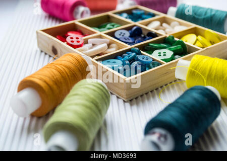 Various colored threads with clothes buttons in wooden box on white cardboard background. Closeup photo. Macro shot Stock Photo