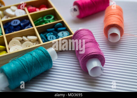 Various colored threads with clothes buttons in wooden box on white cardboard background. Closeup photo. Macro Stock Photo