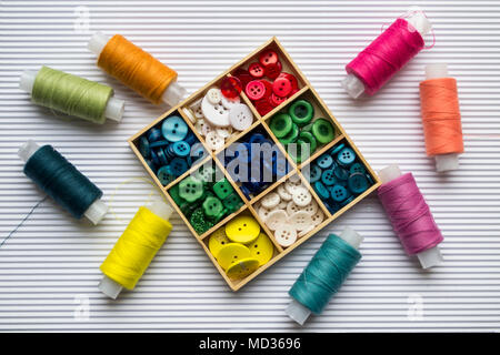 Various colored threads with clothes buttons in wooden box on white cardboard background. Closeup photo. Top view Stock Photo