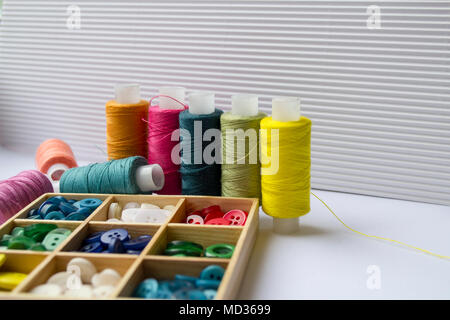 Various colored threads with clothes buttons in wooden box on white cardboard background. Closeup photo Stock Photo