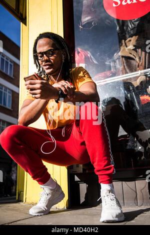 Young man crouching outdoors, wearing earphones, using smartphone Stock Photo