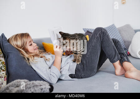 Young girl lying on sofa, reading book, pet cat peering round book Stock Photo