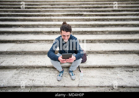 Handsome young man in sports outfit with curly hair sitting on stairs and working on tablet Stock Photo