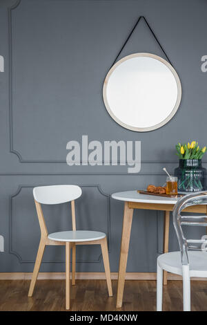 A round mirror with frame on a gray wall with molding above a small, wooden, white dining set in a dark designer interior Stock Photo
