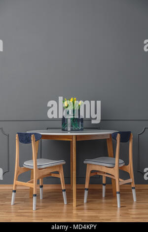 Paint-dipped chairs and a simple, wooden table with a bouquet of yellow tulips in glass vase by an empty, gray wall in a minimalist dining room interi Stock Photo