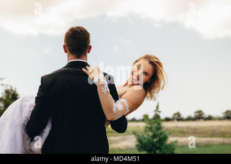 Attractive woman in arms of her husband on wedding day. Rear view of groom carrying smiling bride. Stock Photo