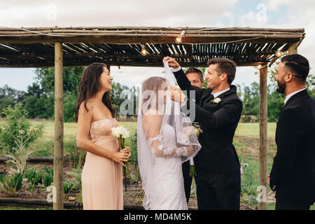 Handsome groom lifting up bridal veil to kiss beautiful bride. Couple having an outdoor wedding ceremony. Bride and groom about to kiss after the wedd Stock Photo