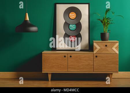 Close-up of a large framed poster with vinyl records leaning against a teal green wall on a modern wooden dresser in a stylish living room interior Stock Photo