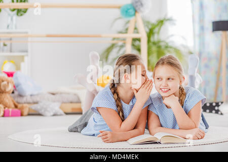 Mischievous twin sisters whispering secrets, lying on a white rug in a cozy children's bedroom Stock Photo