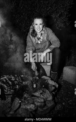 Into the wild. smiling active traveller woman by a bonfire roasting marshmallow Stock Photo