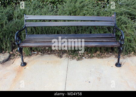 Wooden public bench with black wrought-iron sides on stone sidewalk in front of overgrown hedge with fallen leaves on cold winter day Stock Photo