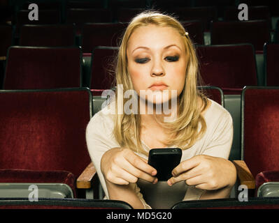 A young woman using her cell phone (iphone) in an old movie theater and not paying attention. Stock Photo