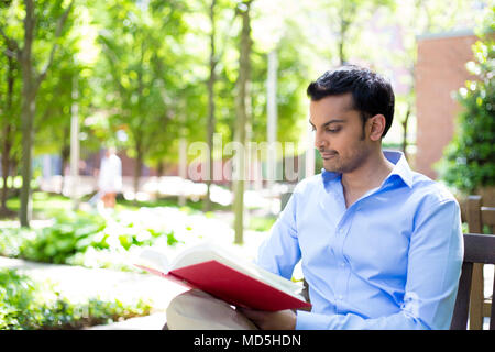 Closeup portrait young business man reading red book, relaxing on wooden bench, sitting outside on sunny day, isolated green trees background Stock Photo