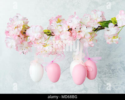 White and pink Easter eggs, decorated with bows, hang on a flowering apple tree branch against a gray stone wall Stock Photo