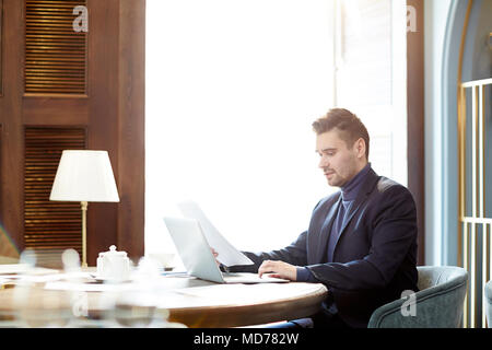 Concentrated middle-aged Caucasian businessman working on laptop and looking through papers in modern office Stock Photo