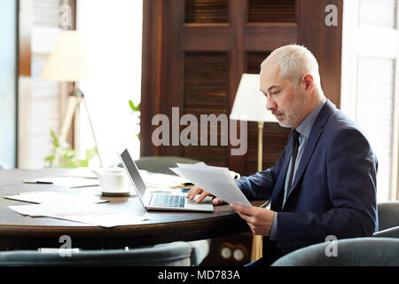 Gray-haired mature businessman working on laptop and looking through documents in modern office Stock Photo