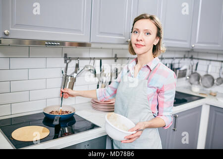Portrait of attractive blond woman standing near electric stove, stirring food in frying pan and smiling at camera Stock Photo