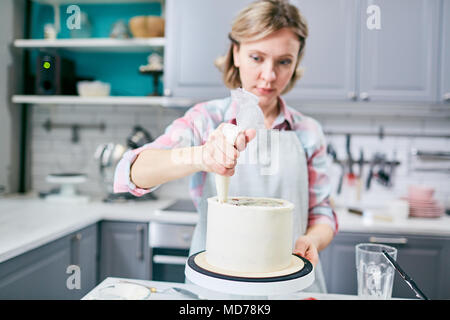Defocused young Caucasian woman using pastry bag while covering appetizing cake with frosting in kitchen Stock Photo