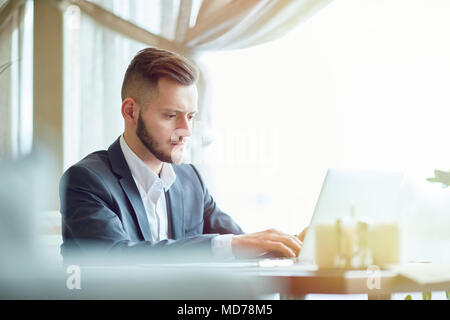 Young handsome Caucasian manager in business attire sitting at cafe table and working on laptop while waiting for lunch Stock Photo