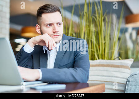 Young good-looking Caucasian male entrepreneur sitting at cafe table with laptop and looking out the window thoughtfully Stock Photo
