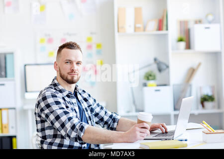 Portrait of young handsome bearded man in checked shirt looking at camera while sitting at laptop with coffee in modern office Stock Photo