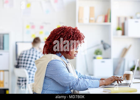 Side view of African American woman with red curly hair sitting at desk and working on laptop in modern office Stock Photo