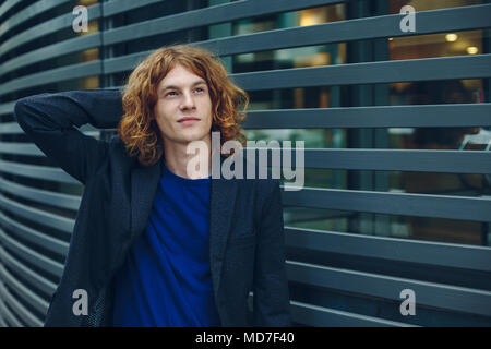 Portrait of young red haired, reddish man with curly hair with arm on head, gray jacket, blue t-shirt and vintage watch over gray urban futuristic bac Stock Photo