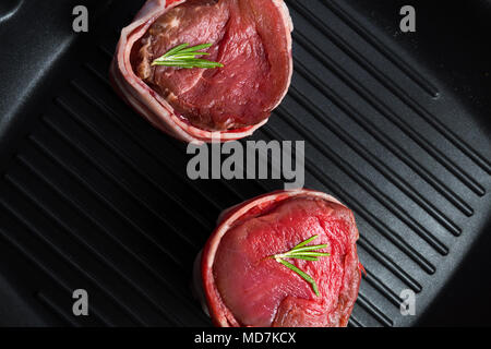 Close-up view from above of two raw beef steaks with bacon in grill pan. Stock Photo