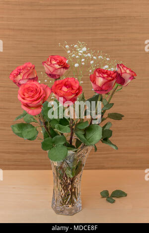 red roses with green leaves in a glass vase with water stand on a wooden table, brown background and blurry white small flowers Stock Photo