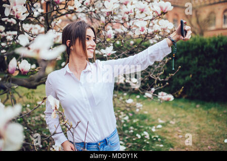 Beautiful stylish caucasian woman making selfie in blossom magnolia garden. Stock Photo