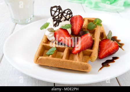 Whole wheat Belgian waffles with whipped cream, freshly chopped strawberries, mint leaves and chocolate on white wooden background. Homemade healthy b Stock Photo