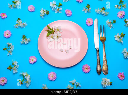 empty round pink ceramic plate and a vintage knife with a fork on a blue surface in the middle of pink and white buds of cherry, top view Stock Photo