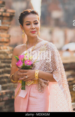 portrait of asian younger woman wearing thai tradition clothes holding pink lotus flower in hand toothy smiling face Stock Photo