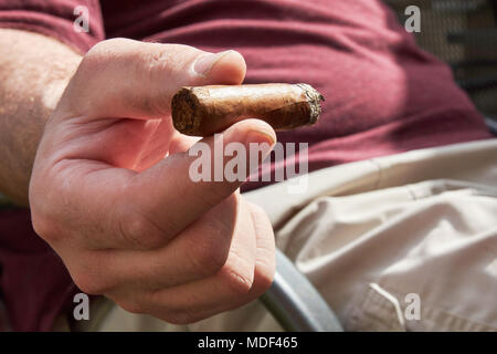 Dirty hand holding a burning cigar, with a blurred man in a red shirt sitting on a chair in the background on a sunny day Stock Photo